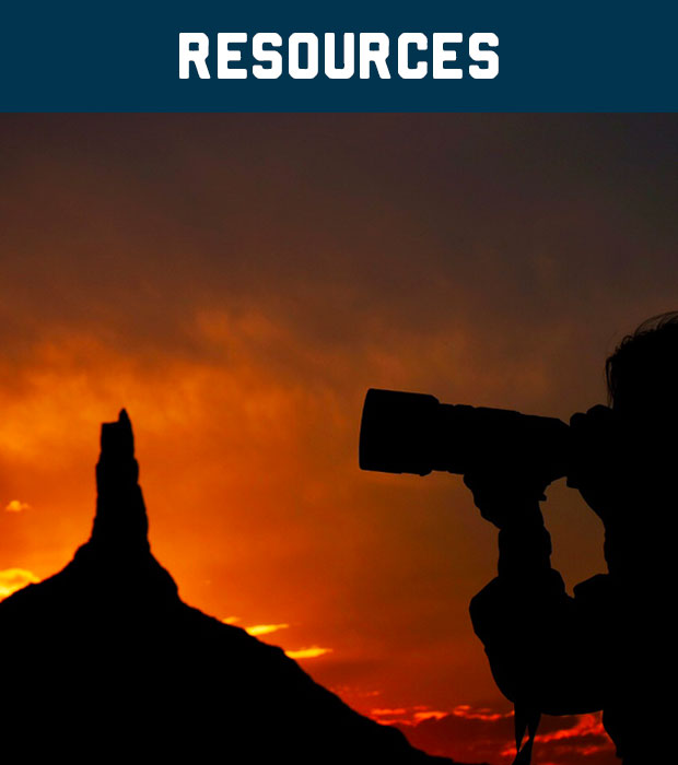 Silhouette of photographer with large lens at sunset with tall rock formation in background.